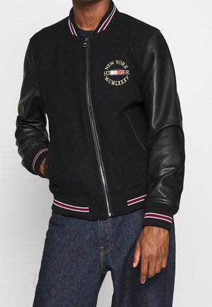 Mand iført sort varsityjakke med ærmer i læder, stribet krave og manchetter, broderet "New York Hilfiger MCMLXXXV" på brystet.