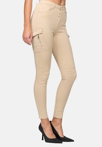 Pantalones cargo beige con bolsillos laterales, corte ajustado y detalle de cremallera. Combinados con zapatos de tacón negro. Textura suave y tejido ligero.