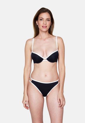Vrouw met lang bruin haar, gekleed in een zwarte bikini met witte rand, staand tegen een effen lichtgrijze achtergrond, voorwaarts gericht.