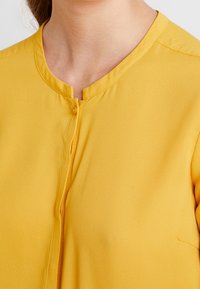 Gelbe Bluse mit rundem Ausschnitt, Knopfverschluss und glatter Textur. Sie hat einen dezenten Glanz, kurze Ärmel und eine Tasche.