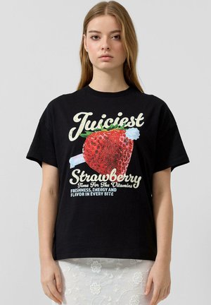 Młoda kobieta ubrana w czarny t-shirt z dużą czerwoną grafiką truskawki i tekstem promującym świeżość, energię oraz witaminy na przodzie.