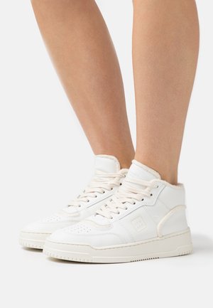 Voeten met witte hoge sneakers met veters, zachte voering en een rubberen zool, afgebeeld tegen een effen witte achtergrond.