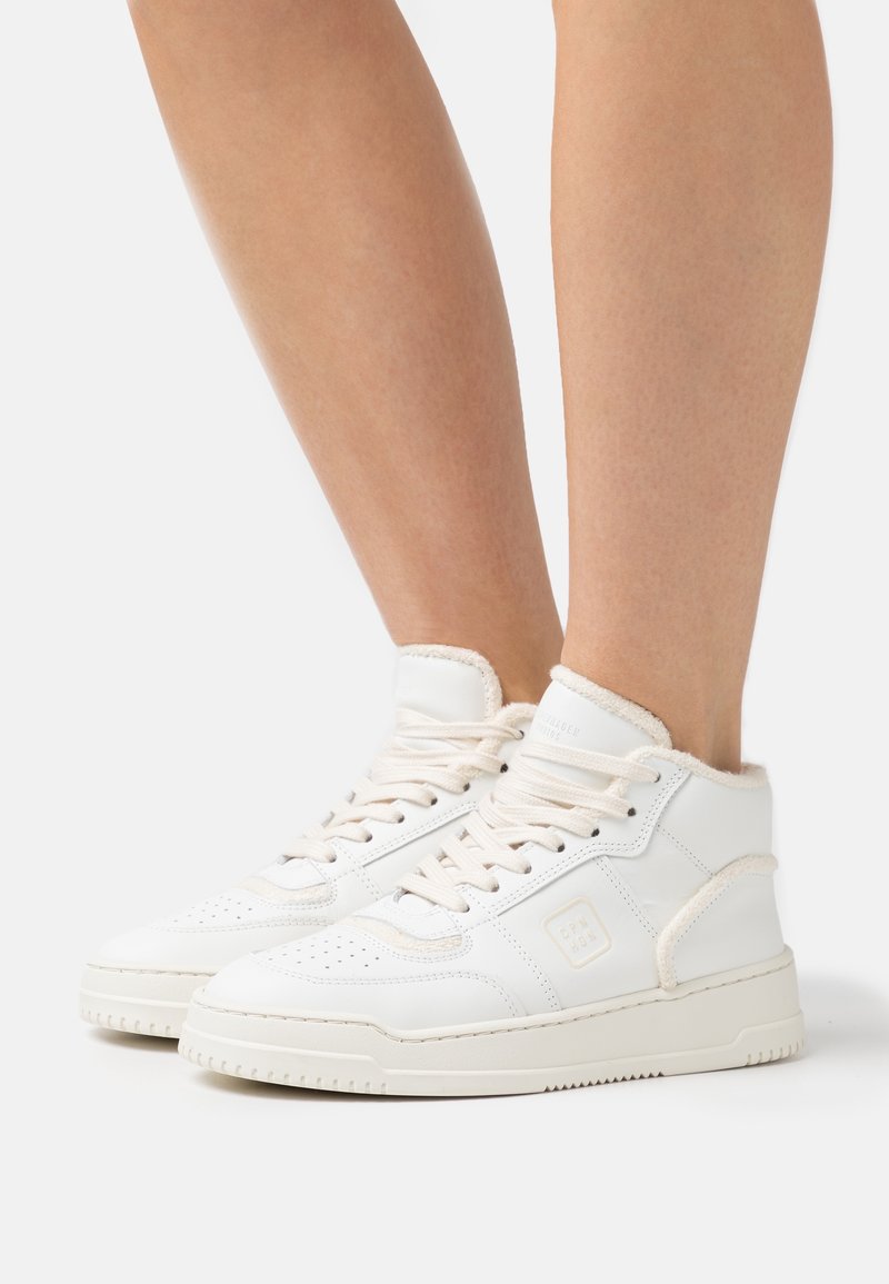 Pieds chaussés de baskets blanches montantes avec lacets, doublure douce et semelle en caoutchouc, présentés sur un fond blanc uni.
