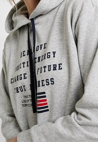 Sweat à capuche gris en tissu doux, avec du texte marine et des rayures rouges, blanches et bleues. Capuche avec cordon de serrage et design graphique à l'avant.