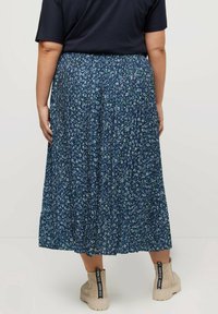 Person med en midikjol i marinblå blommönster, svart topp och beige ankelboots med svarta dragflikar, som står mot en neutral bakgrund.