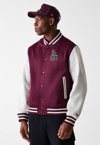 Vinrød varsity-jakke med hvite skinnærmer, ribbede mansjetter og krage. Har "LA"-logo, trykknapper og to sidelommer.