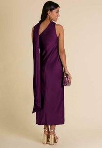 Paarse satijnen jurk met een gedrapeerde achterkant en een zijsplit, met een zachte textuur. Gecombineerd met een doorzichtige clutch en metallic platformhakken.