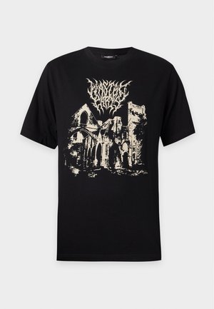 Junger Mann trägt ein schwarzes T-Shirt mit einer weißen Grafik eines zerstörten Gebäudes und abstraktem Text auf der Brust, steht vor einem schlichten Hintergrund.