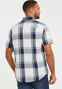Camisa de cuadros de manga corta en gris y azul marino con detalles en blanco. Cuenta con un cuello, cierre de botones en la parte posterior y un dobladillo ligeramente curvado.