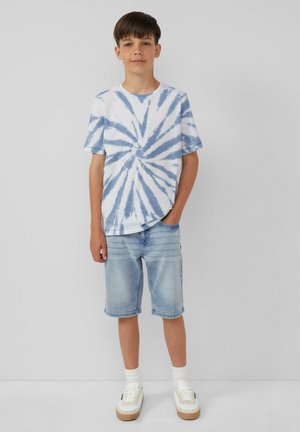 Blau-weißes Batik-T-Shirt mit kurzen Ärmeln, kombiniert mit hellblauen Denim-Shorts und weißen Sneakers mit beigen Sohlen.