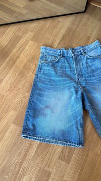 Shorts en denim bleu clair avec une coupe décontractée, dotés d'un design à cinq poches, d'un bouton supérieur et d'une texture effet dégradé. Posés à plat sur un sol en bois.