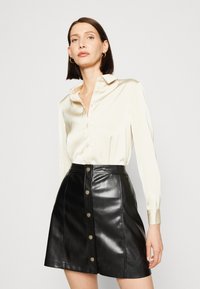 Kremo satino palaidinė su apykakle, užsagstoma. Juoda odinė A formos sijonas su šešiais auksinės spalvos mygtukais priekyje. Abu daiktai turi lygią tekstūrą.