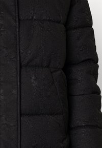 Veste matelassée noire à motifs cousus avec poche latérale, montrant la partie centrale du corps et une manche.