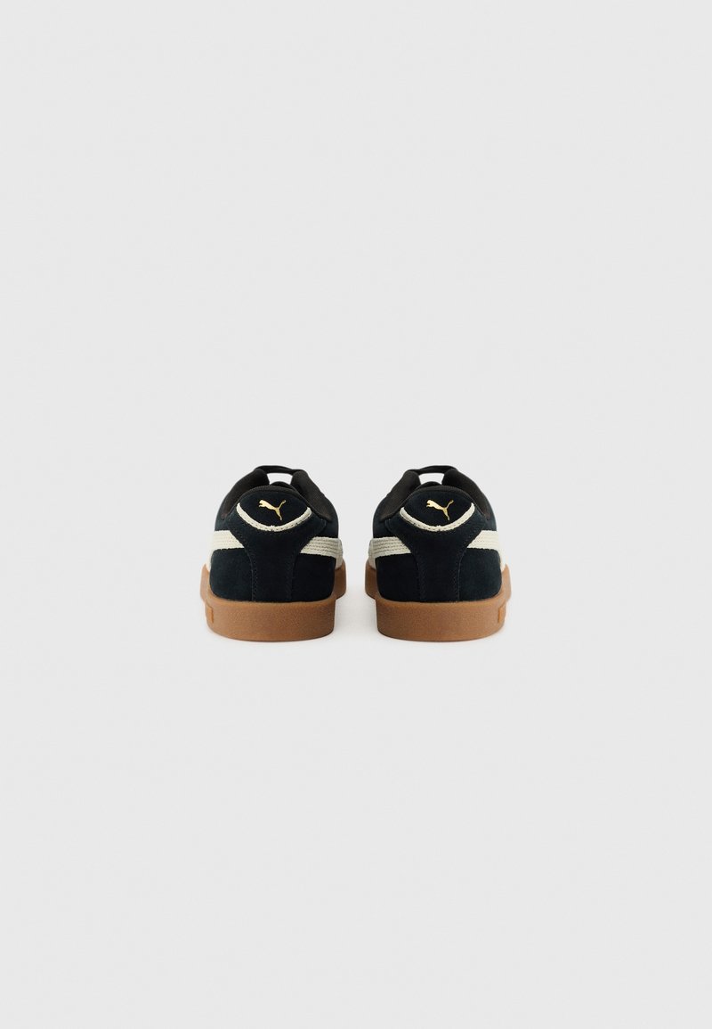 Sneakers neri in suede con suola in gomma beige, caratterizzati da una striscia bianca e un logo dorato sul tallone. Design semplice, forma a taglio basso.