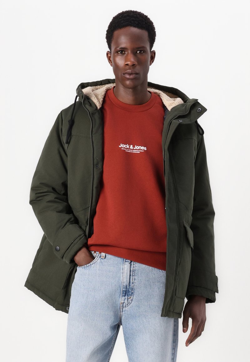 Parka verte avec une capuche doublée de fourrure, portée sur un sweat-shirt rouge avec du texte blanc, associée à un jean en denim clair.
