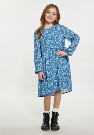 Blå floral kjole med lange ærmer og samlet talje, prydet med hvide margueritter og et let stof. Sorte støvler med snørebånd fuldender outfittet.