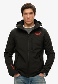Zwarte, met rits sluitende jas met een capuchon; heeft rode logo-accenten, een gladde textuur en zijzakken. Draag het over een grijze sweater met een spijkerbroek.