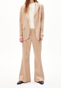 Costume beige ajusté avec un blazer structuré et un pantalon évasé, associé à un haut blanc. Présente des lignes épurées et des revers nets. Chaussures noires à talons pointus.