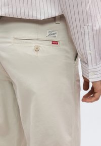 Μπεζ πίσω τσέπη παντελονιού Levi's με κουμπί και κόκκινη ετικέτα Levi's, φορεμένο με λευκό πουκάμισο με λεπτές μαύρες κάθετες ρίγες.