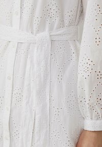 Vestito bianco in cotone con dettagli in pizzo, vita con cintura e chiusura a bottoni. Presenta maniche lunghe con orli a risvolto e motivi floreali.