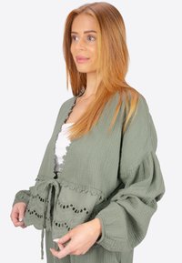 Hellgrüne, kurze Bluse aus strukturiertem Stoff mit Spitzenbesatz und gebundenem Vorderteil. Lässige Ärmel mit gerafften Bündchen, die einen entspannten Schnitt betonen.