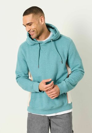 Mann lächelt und schaut nach unten, trägt einen türkisfarbenen Kapuzenpullover mit Kordelzügen und graue Hose vor einem schlichten hellen Hintergrund.