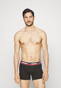 Fekete pamut boxer alsónadrág fehér, piros és fekete derékrésszel, amelyen a "LEVI'S" felirat szerepel, a modell ing nélkül áll egy világos háttér előtt.