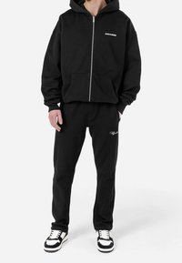 Felpa nera con zip e tasche frontali, con la scritta bianca "PEGADOR". Pantaloni in cotone a coste neri coordinati con dettagli del logo bianco. Sneakers in bianco e nero.
