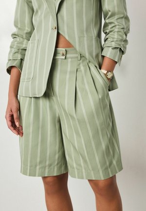 Woman wears light green pinstripe blazer, matching pleated shorts, and a wristwatch, with one hand in pocket against plain background.