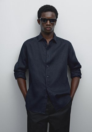 Dark blue, button-up shirt with rolled sleeves, paired with black sunglasses. Smooth texture against a neutral background.
