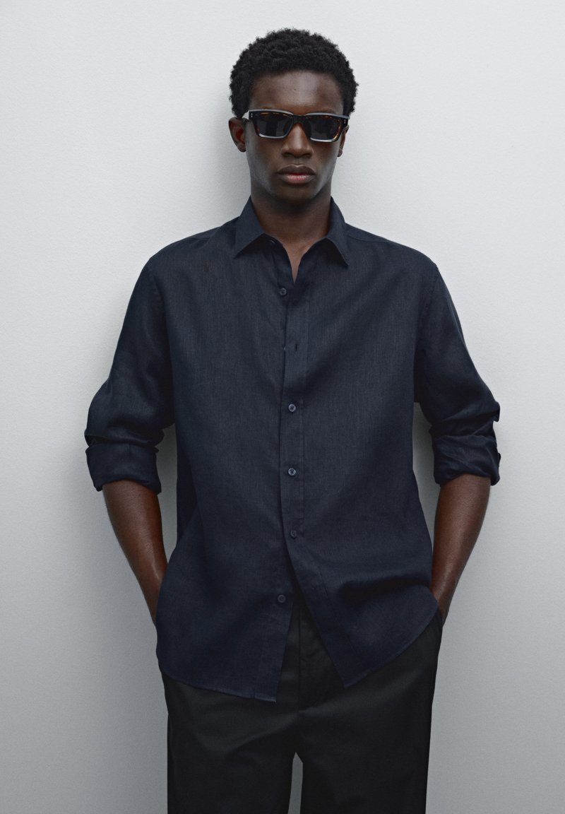 Dark blue, button-up shirt with rolled sleeves, paired with black sunglasses. Smooth texture against a neutral background.
