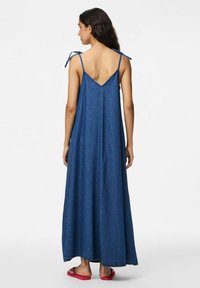 Duga, bezrukava traper haljina tamnoplave boje, s V-izrezom na leđima, podesivim trakama za vezanje i lepršavim suknjom. Uparena s ravnim ružičastim sandalama.