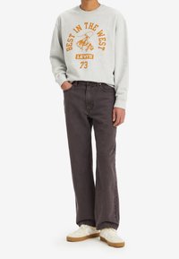 Felpa grigia con testo e logo arancioni, abbinata a jeans grigio scuro. Le sneakers presentano un design bianco e crema con suola marrone chiaro.