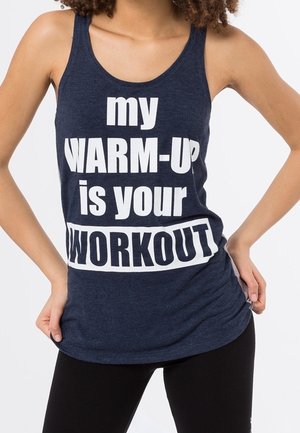 Femme portant un débardeur bleu marine avec un texte blanc en gras indiquant « mon ÉCHAUFFEMENT est votre ENTRAÎNEMENT », posant les mains sur les hanches.