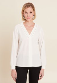 Weiße Bluse mit V-Ausschnitt, langen Ärmeln und strukturiertem Stoff. Verfügt über eine dezente zentrale Naht und geknöpfte Ärmel. Kombiniert mit schwarzen Hosen.