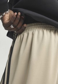 Main à la peau foncée avec des bagues et un bracelet en argent glissés dans une taille élastique de pantalon beige à rayures verticales noires, portant un haut noir.