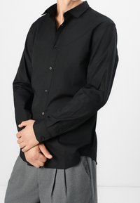Schwarzes Langarm-Hemd mit Knopfleiste und weitem Kragen, aus glattem Stoff mit glänzenden Knöpfen, kombiniert mit grauen, gefalteten Hosen.
