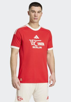 Mann trägt rotes 1. FC Union Berlin Fußballtrikot und weiße Shorts, steht vor hellem, einfarbigem Hintergrund, leicht nach links gewandt.