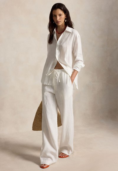 Γυναίκα που φοράει λευκό λινό πουκάμισο και παντελόνι, χαλαρής γραμμής, με κόκκινο κολιέ και σκουλαρίκια, κρατώντας ένα πλεγμένο τσάντα, στέκεται σε ουδέτερο φόντο.