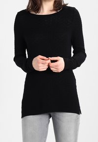 Woman wearing a textured black long-sleeve top with side slits, paired with light gray jeans, hands clasped at waist.