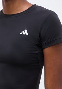 Κοντινό πλάνο ατόμου που φοράει εφαρμοστή μαύρη αθλητική κοντομάνικη μπλούζα με μικρό λευκό τρίγωνο λογότυπο στο αριστερό στήθος.