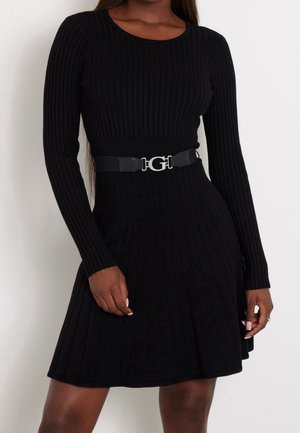 Femme portant une robe noire en maille côtelée à manches longues avec une ceinture noire ornée d'une boucle argentée "G", debout devant un fond uni.