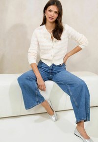 White cardigan with textured design, light pastel accents, paired with wide-leg blue denim jeans and shiny silver ballet flats.