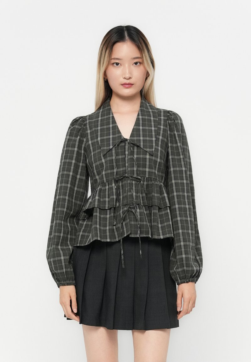 Dark plaid blouse with puffed sleeves, peplum waist, and drawstring detail, paired with a black pleated skirt.