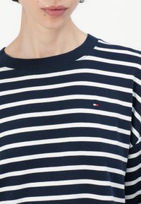 Camisa de algodón a rayas en azul marino y blanco con cuello redondo y mangas cortas; presenta un pequeño logo en rojo, blanco y azul en el pecho.