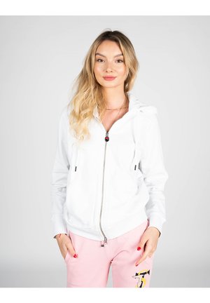 Felpa bianca con cerniera e cappuccio, due tasche frontali e cordini neri; indossata con pantaloni rosa chiaro con un accento ricamato.