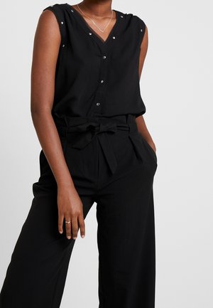 Woman wearing sleeveless black buttoned V-neck top and high-waisted black pants with tied belt, hand in pocket against plain background.