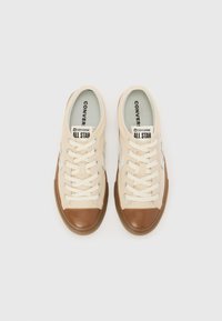 Beige lerretsneakers med en brun gummitåhett og teksturert såle, med hvite snøring og en lys blå innside. "Converse All Star"-merket synlig.