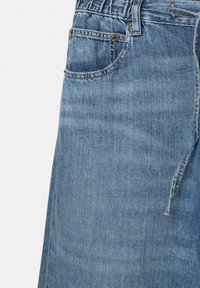Gros plan sur un jean en denim bleu montrant la poche avant, les passants de ceinture et les détails visibles de couture.