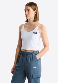 Jeune femme portant un crop top blanc et un pantalon bleu à taille élastique, tous deux avec le logo The North Face, debout avec une main dans la poche.